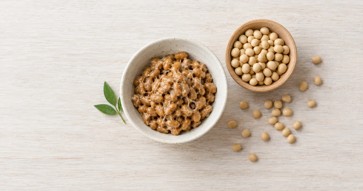 Natto em uma tigela clara, ao lado de uma tigela de madeira e de grãos de soja soltos sobre superfície clara de madeira